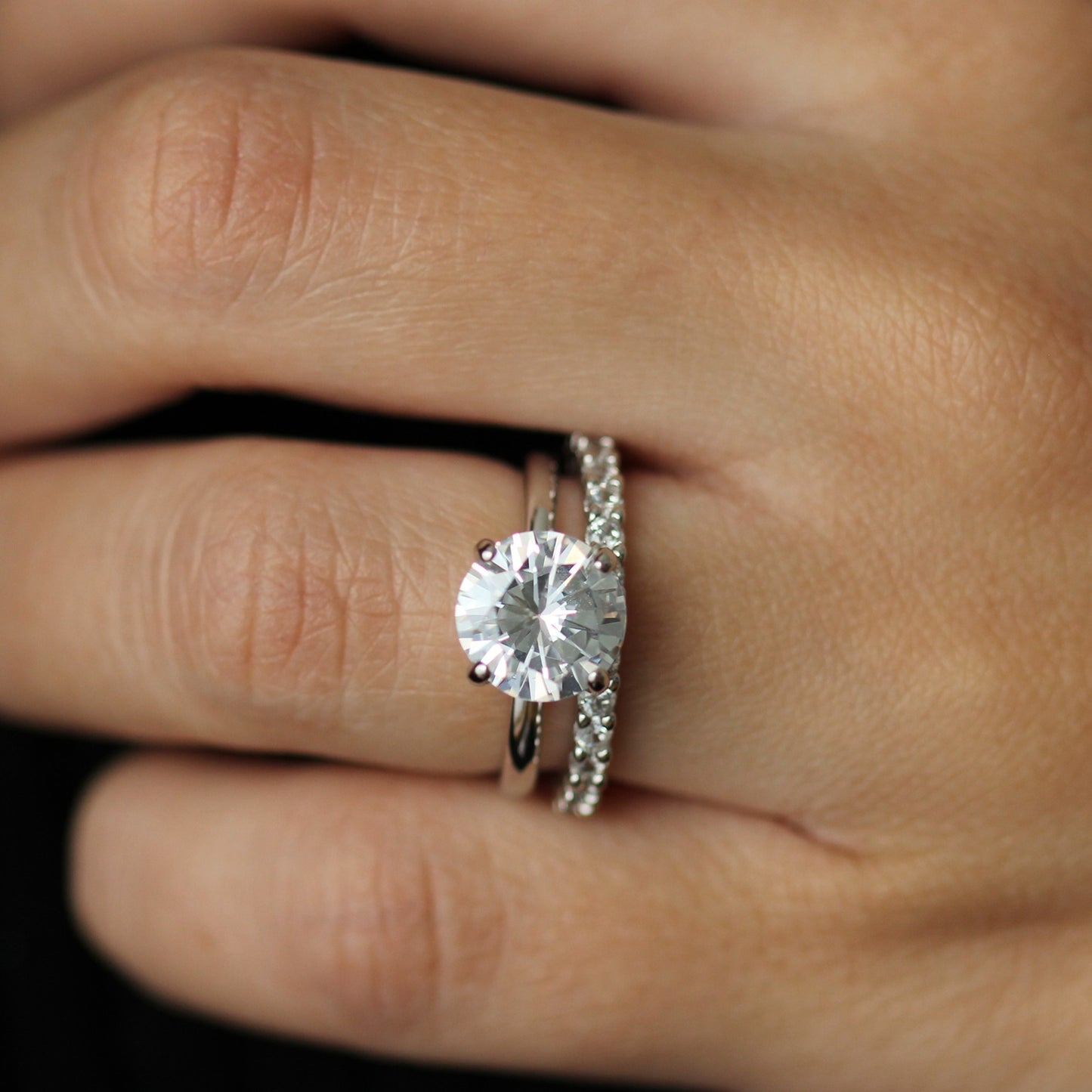 Close-up of a hand wearing a 3 ct round cut ring and eternity band with a dark background